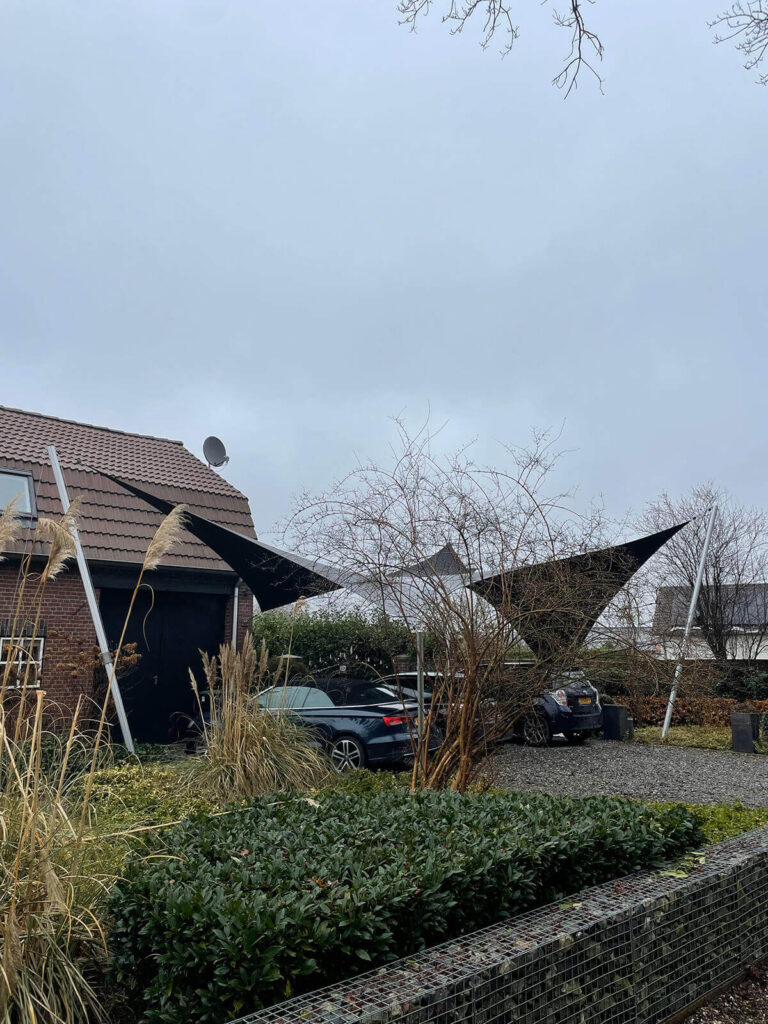 Black triangular shade sails over a driveway with cars, a brick house, and bare trees on a cloudy day.