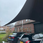 Shade sail stretched across a driveway above parked cars beside brick houses on a cloudy day