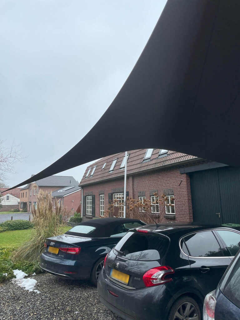 Shade sail stretched across a driveway above parked cars beside brick houses on a cloudy day
