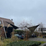Two black triangular shade sails stretch over a driveway in a suburban front yard, with parked cars, bare trees, and a cloudy sky.