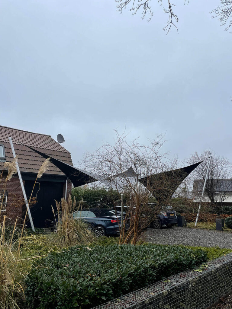 Two black triangular shade sails stretch over a driveway in a suburban front yard, with parked cars, bare trees, and a cloudy sky.