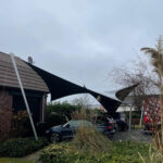 Black sunshade sail stretched between a brick house and poles, covering a row of parked cars in a suburban driveway.