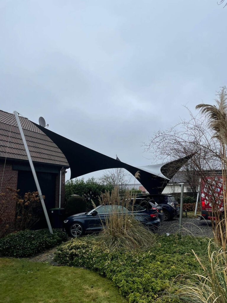 Black sunshade sail stretched between a brick house and poles, covering a row of parked cars in a suburban driveway.