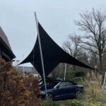 Large black fabric shade sail stretched between metal poles over a parked blue car in a residential yard.