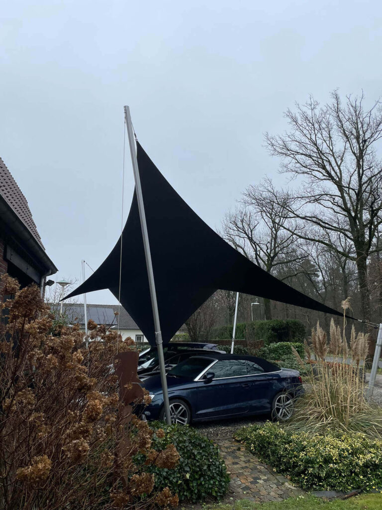 Large black fabric shade sail stretched between metal poles over a parked blue car in a residential yard.