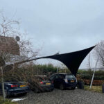 Black fabric shade sails stretched between metal poles over a car park on a cloudy day, with several cars parked nearby.