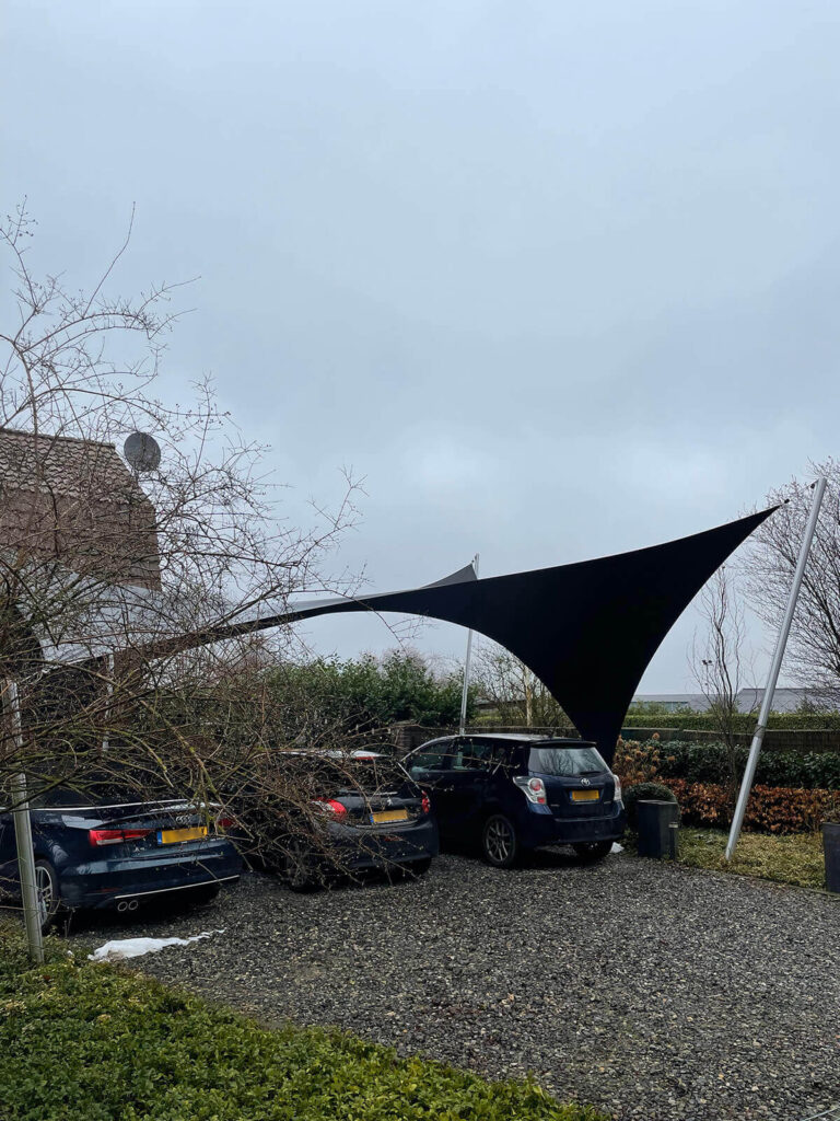 Black fabric shade sails stretched between metal poles over a car park on a cloudy day, with several cars parked nearby.