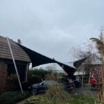 Black tensile shade sail spans between a brick house and a pole, shading a blue car in the driveway under a cloudy sky.