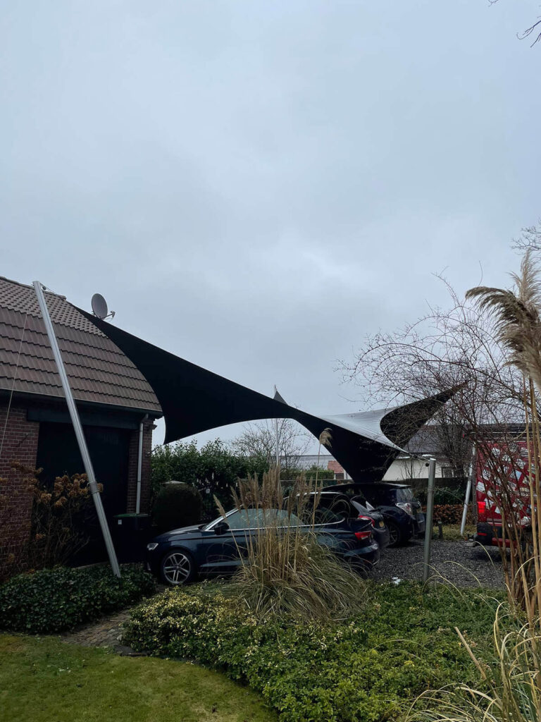 Black tensile shade sail spans between a brick house and a pole, shading a blue car in the driveway under a cloudy sky.