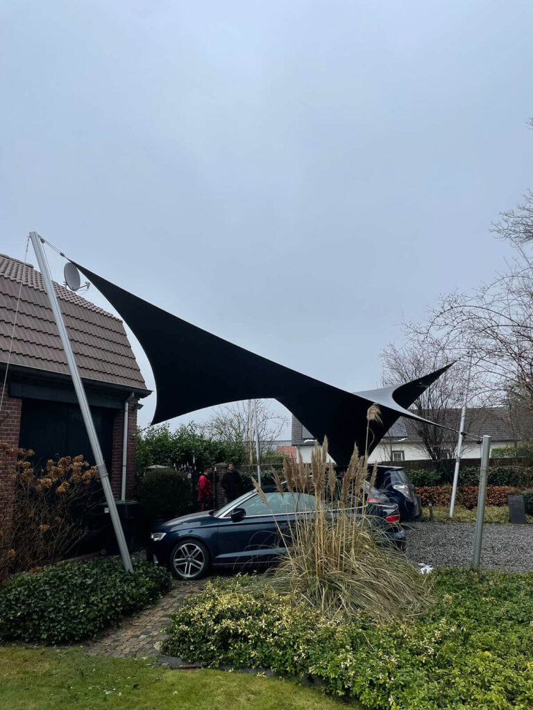 Black sail shade canopy stretched between metal poles over a driveway with a blue car parked nearby and a brick house in the background