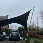 Large black shade sail stretched over a residential driveway, supported by a tall pole, with cars parked below.