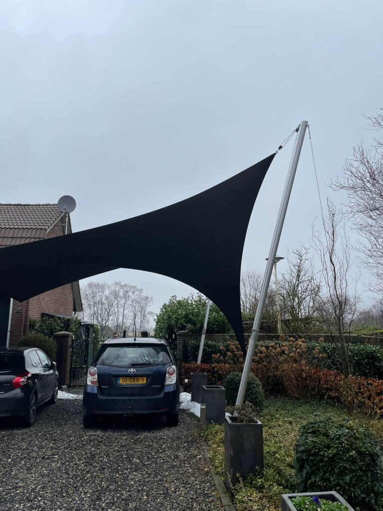 Large black shade sail stretched over a residential driveway, supported by a tall pole, with cars parked below.