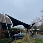 Large black shade sail stretched between metal poles over a driveway with a blue car parked beneath; two people stand nearby by the hedge.