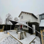 Snowy backyard with a modern patio, a large triangular black shade sail, string lights along the house, and a person in red near the door.