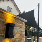 Stone wall with warm yellow uplighting and a black modern wall light, string lights along a building, and an outdoor dining area with chairs in the background.