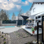 Backyard with a rectangular pool, stone patio, and modern seating area under a shade sail by a white house.