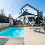 Backyard with a rectangular pool, wooden deck, and a white house with a black shade sail overhead.