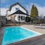 Backyard of a modern white house with a rectangular pool, wood deck, and a black shade sail over outdoor seating.