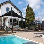 Modern white two-story house with a black shade sail over the patio by a pool; a small dog in the foreground.