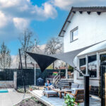 Modern backyard with white brick house, a black shade sail, and a wooden outdoor seating area by a poolside patio.