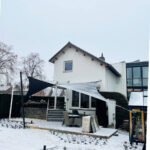 White two-story house with a snow-covered yard and a black triangular shade sail over the patio furniture.