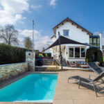 Backyard scene with a rectangular pool, tan patio, black shade sail, white two‑story house, outdoor seating, and a greenhouse on the right.