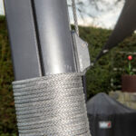 Close-up of a braided steel cable wrapped around a dark gray metal post, secured by a clamp with a small screw visible (outdoor setting).