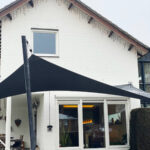 White brick house with large glass doors, string lights along the roof, and a black shade sail over an outdoor seating area.