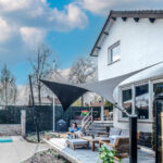 Backyard patio with a white brick house, pool, and a large triangular shade sail over a seating area.