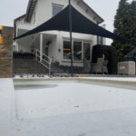 White two-story house with string lights along the eaves, a large black shade sail over a patio, and snow-covered ground; outdoor dining chairs nearby with a small dog on the steps as a backdrop.