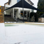 White two-story house with string lights along the eaves, a large black fabric shade sail over a patio, and a snow-covered yard.