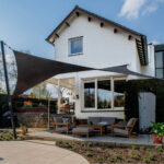 Modern white house with large glass patio doors and a black shade sail over the outdoor seating area.