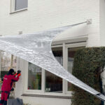 Snow-covered curved metal awning attached to a white brick house, with a worker in a red jacket nearby next to a window.