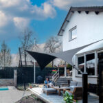 Backyard patio scene with a white brick house, modern seating under a black and gray shade sail, and a pool nearby.
