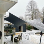 Snowy backyard with a large black shade sail attached to a white house and glass conservatory below the sail-ground covered in light snow.