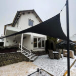 White two-story house with string lights along the roof, and a large black triangular shade sail over a snowy outdoor dining area.