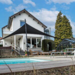 White two-story house with a black shade sail over a patio seating area beside a pool; a dog nearby.