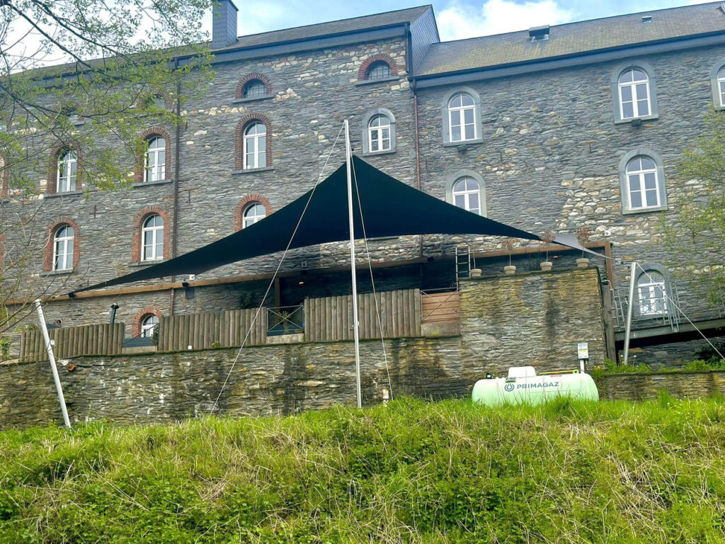 Black triangular shade sail stretched over an outdoor seating area beside a gray stone building with arched windows.