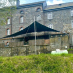 Black triangular shade sail stretched over an outdoor seating area beside a gray stone building with arched windows.