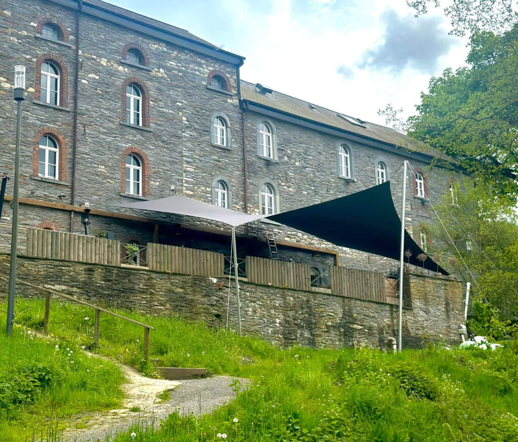 Stone building with arched windows on a grassy hillside and wooden fencing below the deck area with shade sails overhead.