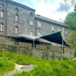 Stone building with arched windows on a grassy hillside and wooden fencing below the deck area with shade sails overhead.
