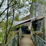 Wooden walkway with metal railings along a stone building, shaded by tree branches, leading to a courtyard with people and seating.