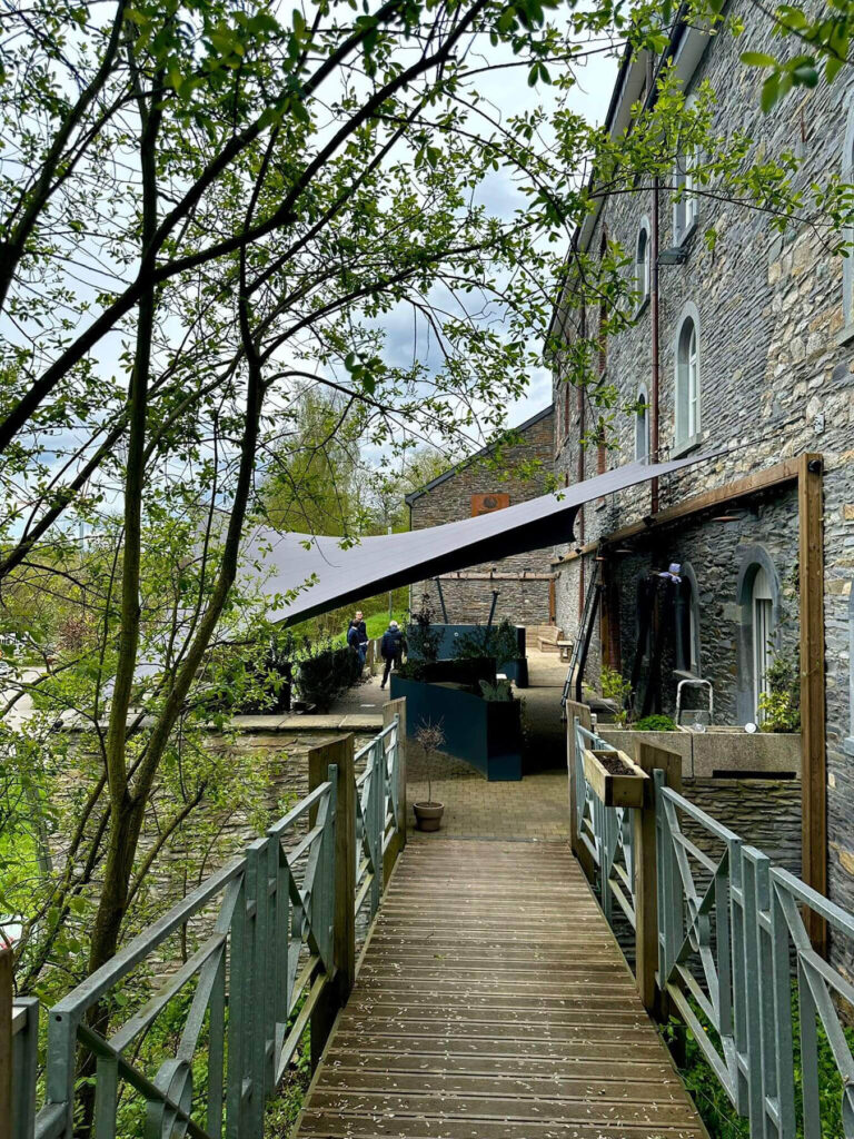 Wooden walkway with metal railings along a stone building, shaded by tree branches, leading to a courtyard with people and seating.