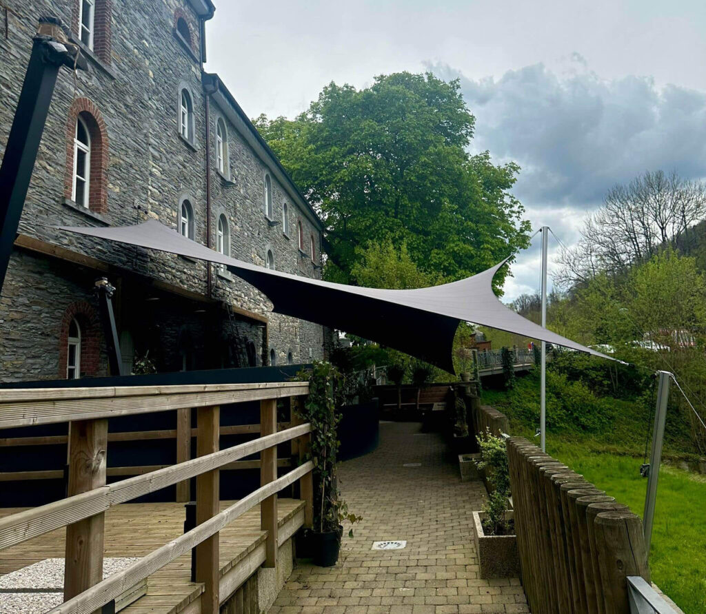 Stone building with arched windows, a wooden railing, and a large fabric shade sail stretched over an outdoor walkway.
