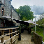 Stone building with arched windows, a wooden railing, and a large fabric shade sail stretched over an outdoor walkway.
