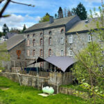 Stone building with arched windows and slate roof, outdoor seating area under a dark canopy, green lawn in foreground