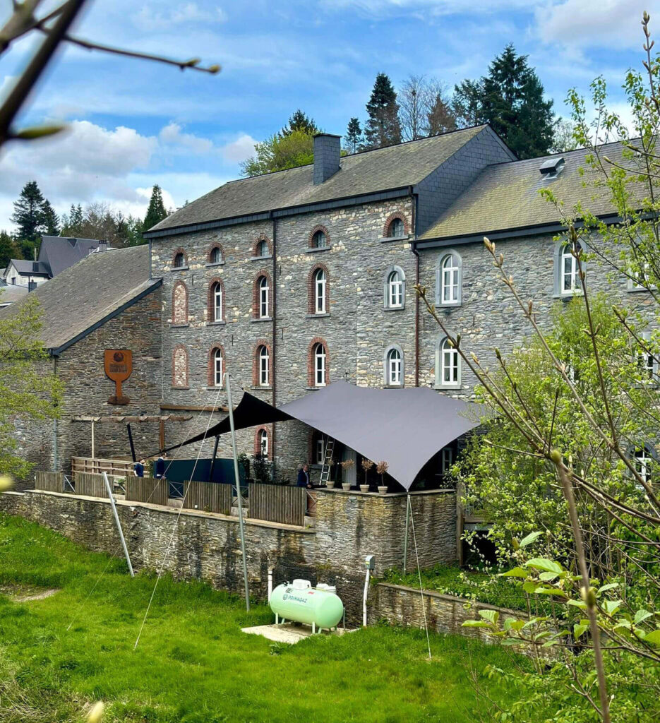 Stone building with arched windows and slate roof, outdoor seating area under a dark canopy, green lawn in foreground
