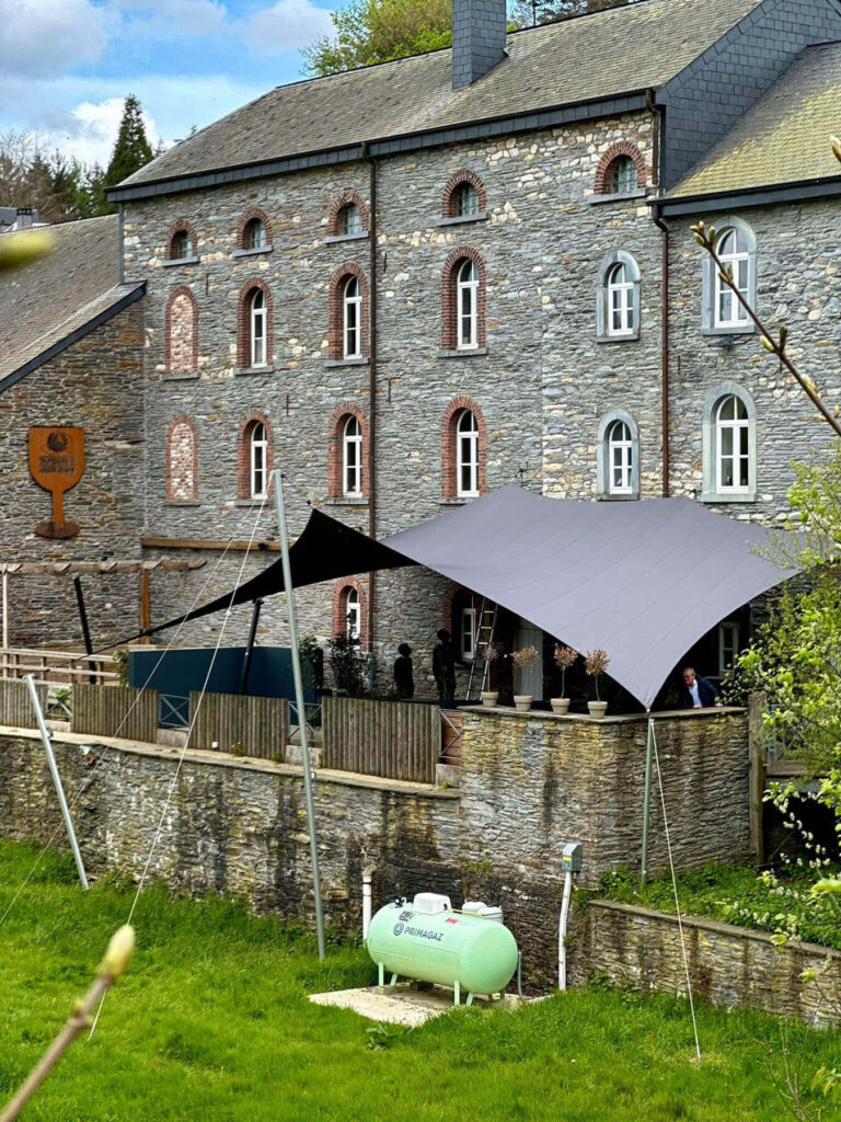 Stone building with arched windows and a large gray canopy over an outdoor area; a Primagaz propane tank nearby on the wall.