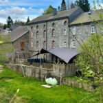 Stone building with arched windows and slate roof, outdoor seating under a large gray shade sail.