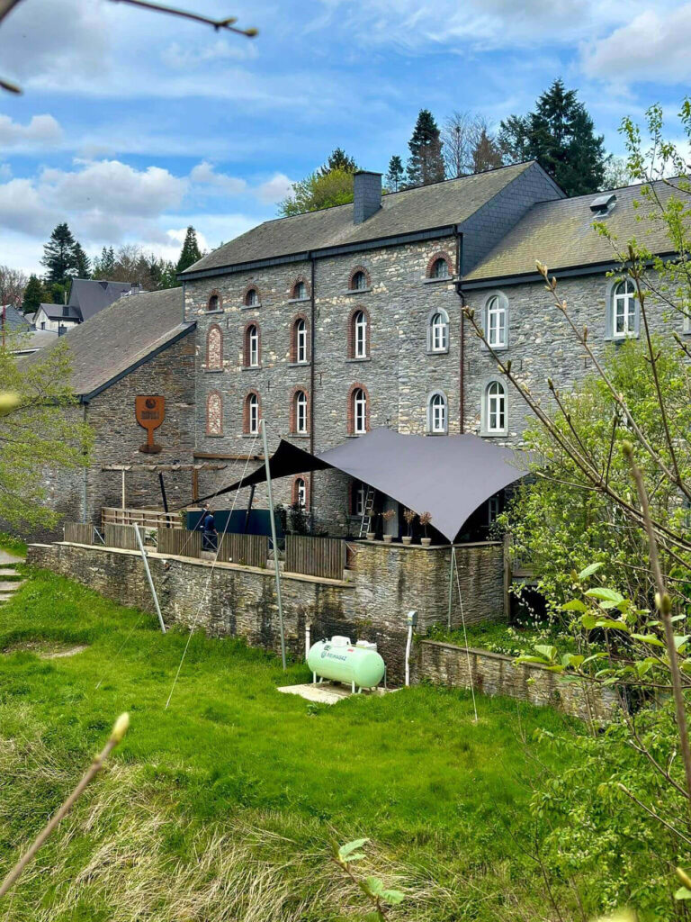 Stone building with arched windows and slate roof, outdoor seating under a large gray shade sail.